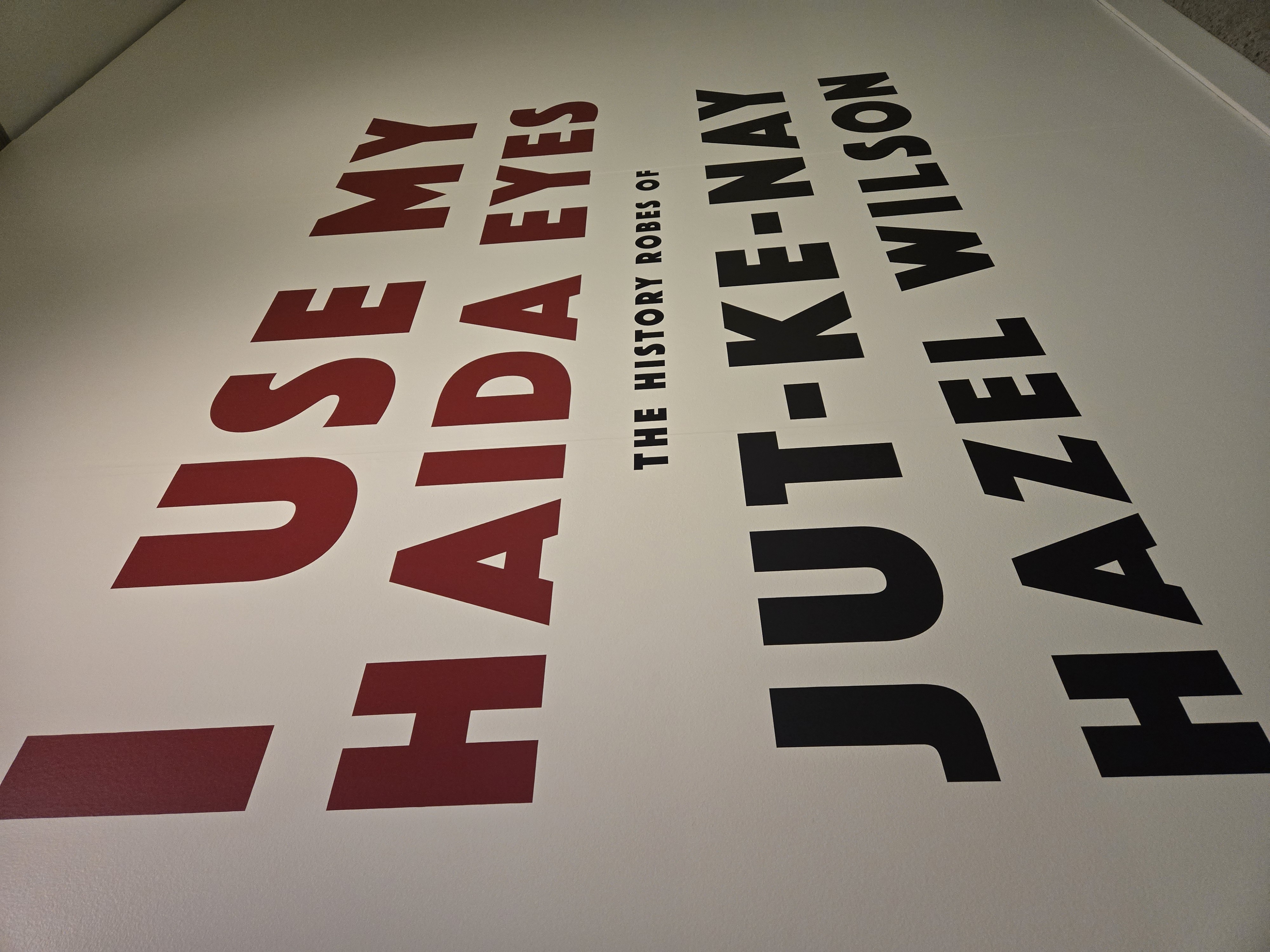 Large red and black wall text reading “I Use My Haida Eyes – The History Robes of Jut-ke-Nay Hazel Wilson” displayed in a gallery exhibition space.