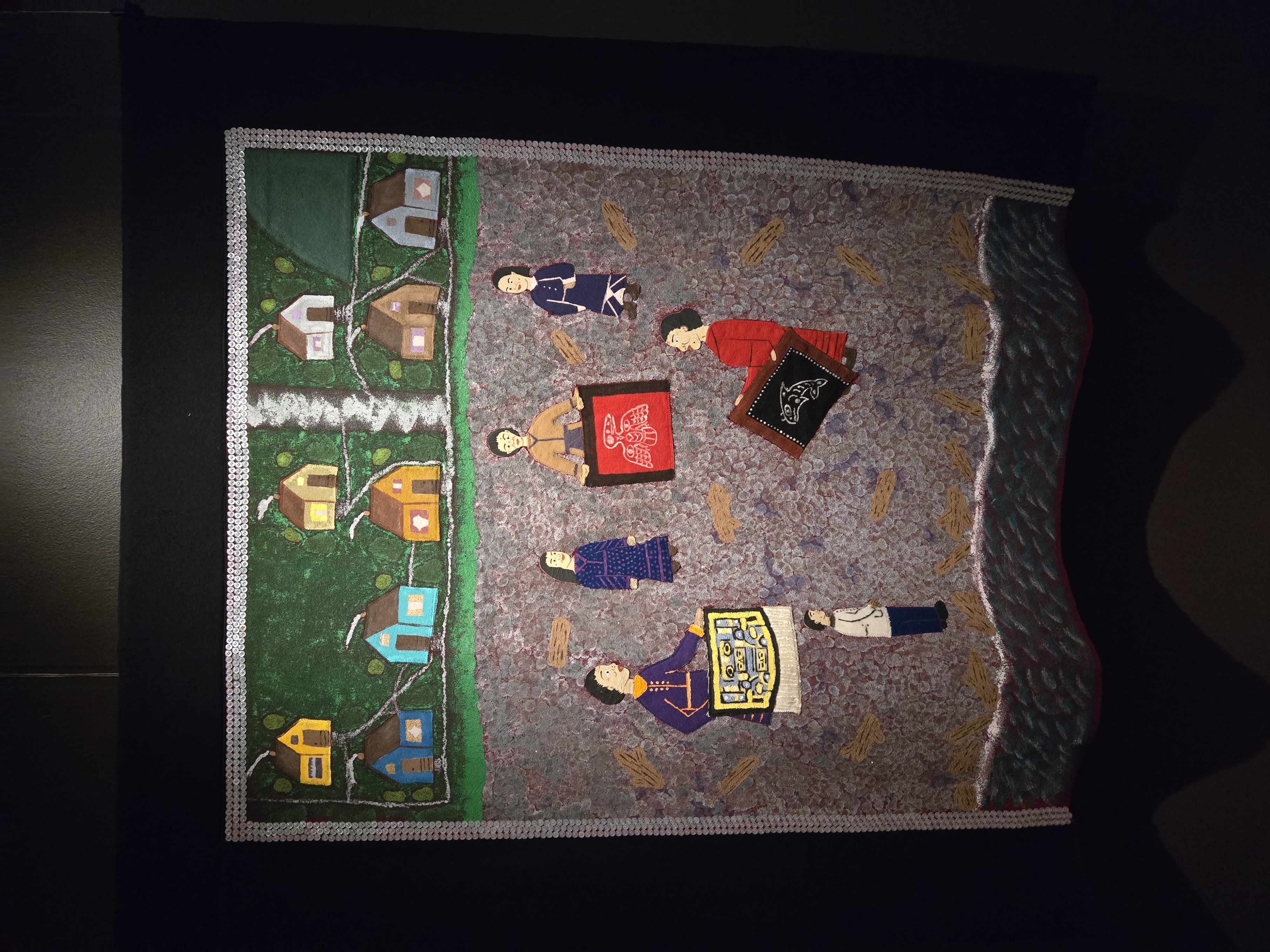 Story robe artwork showing small houses connected by paths above figures gathered around ceremonial objects, illustrating community life and cultural storytelling.