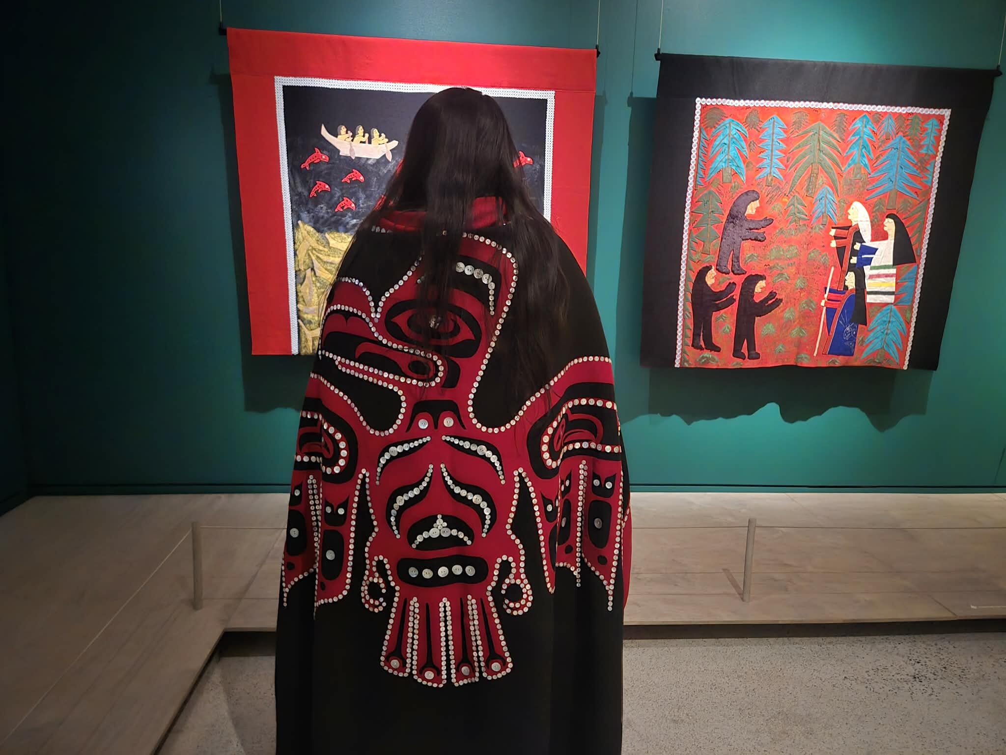 Visitor wearing a red and black Haida-style robe with formline design stands in a gallery, viewing two story robes depicting figures, animals, and landscapes on teal walls.