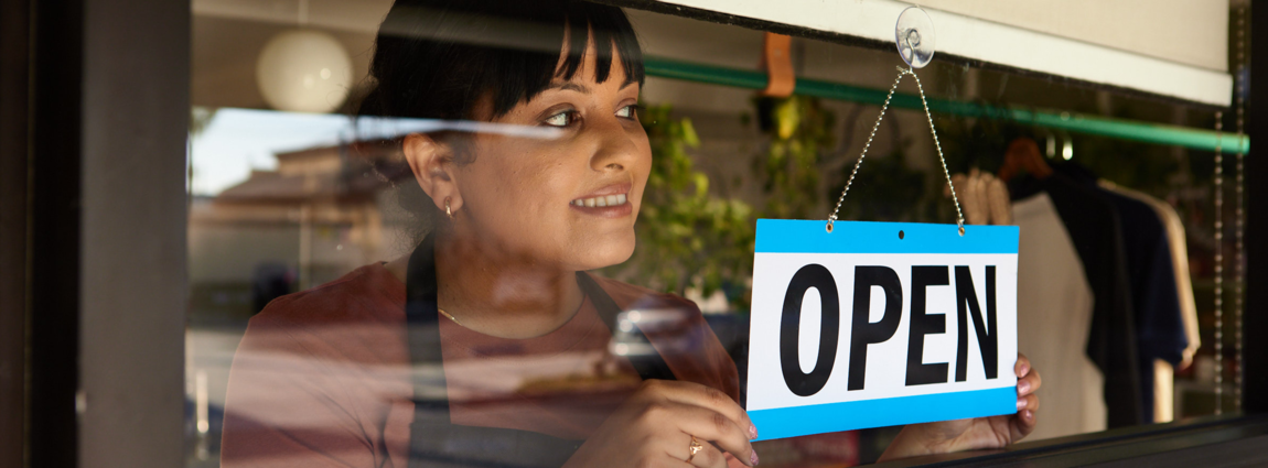 Woman wearing an apron and brown tshirt behind a glassdoor and open sign