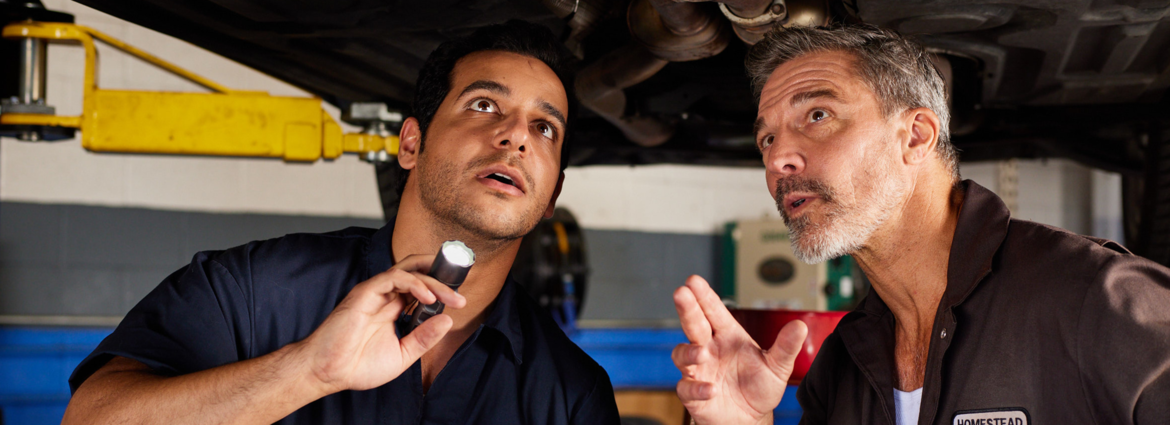 Two men standing under a vehicle on a lift. One man holding a flashlight, the other man is pointing at the underside of the vehicle